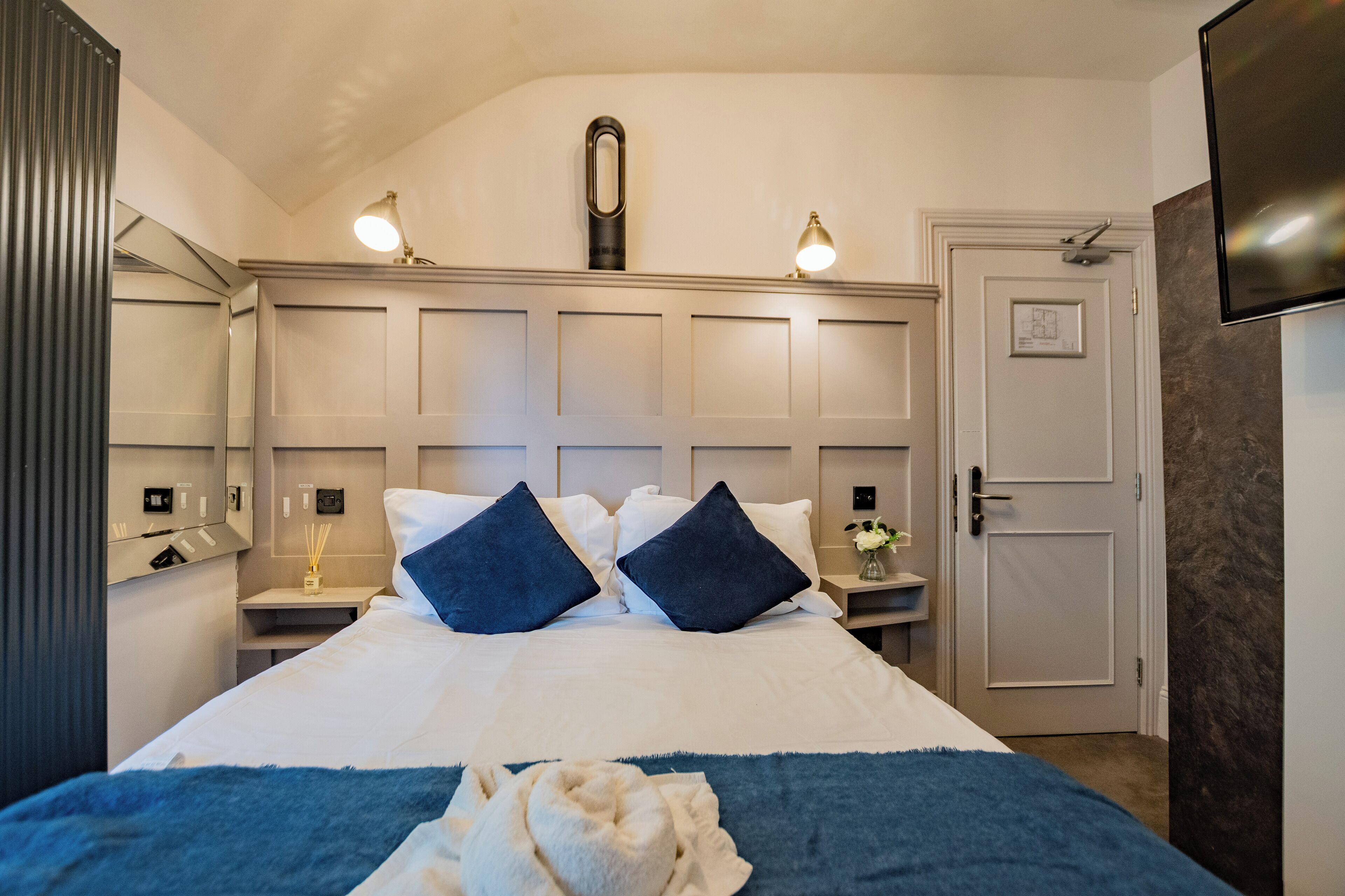 Ein ordentlich eingerichtetes Hotelzimmer mit einem großen Bett, zwei Nachttischen, einem Spiegel und einer Tür mit einem gerahmten Bild.