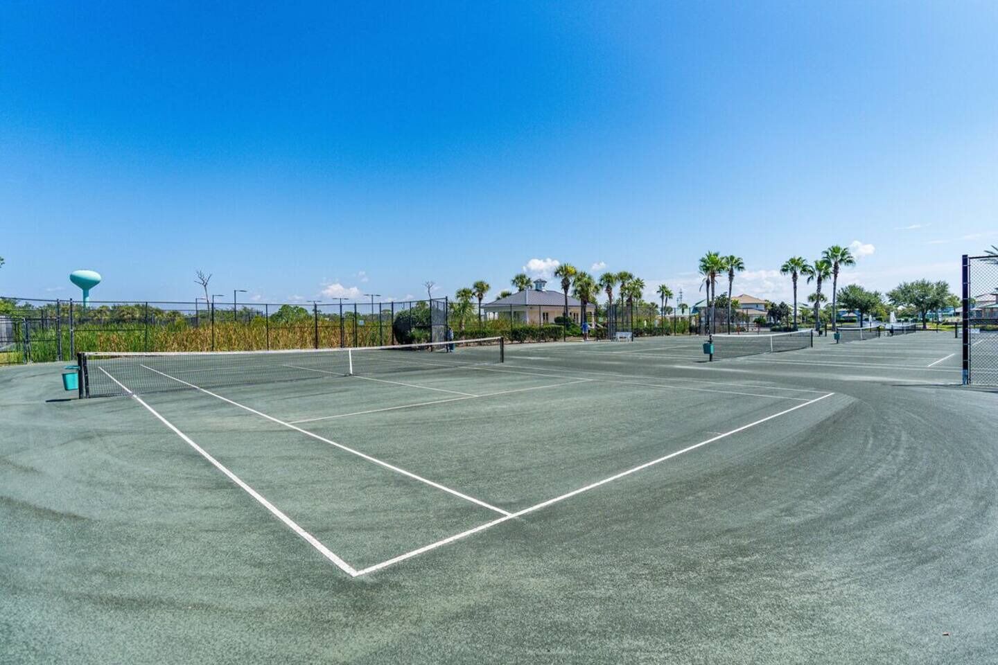 A tennis court with a net, surrounded by a fence, palm trees, and buildings in the background.