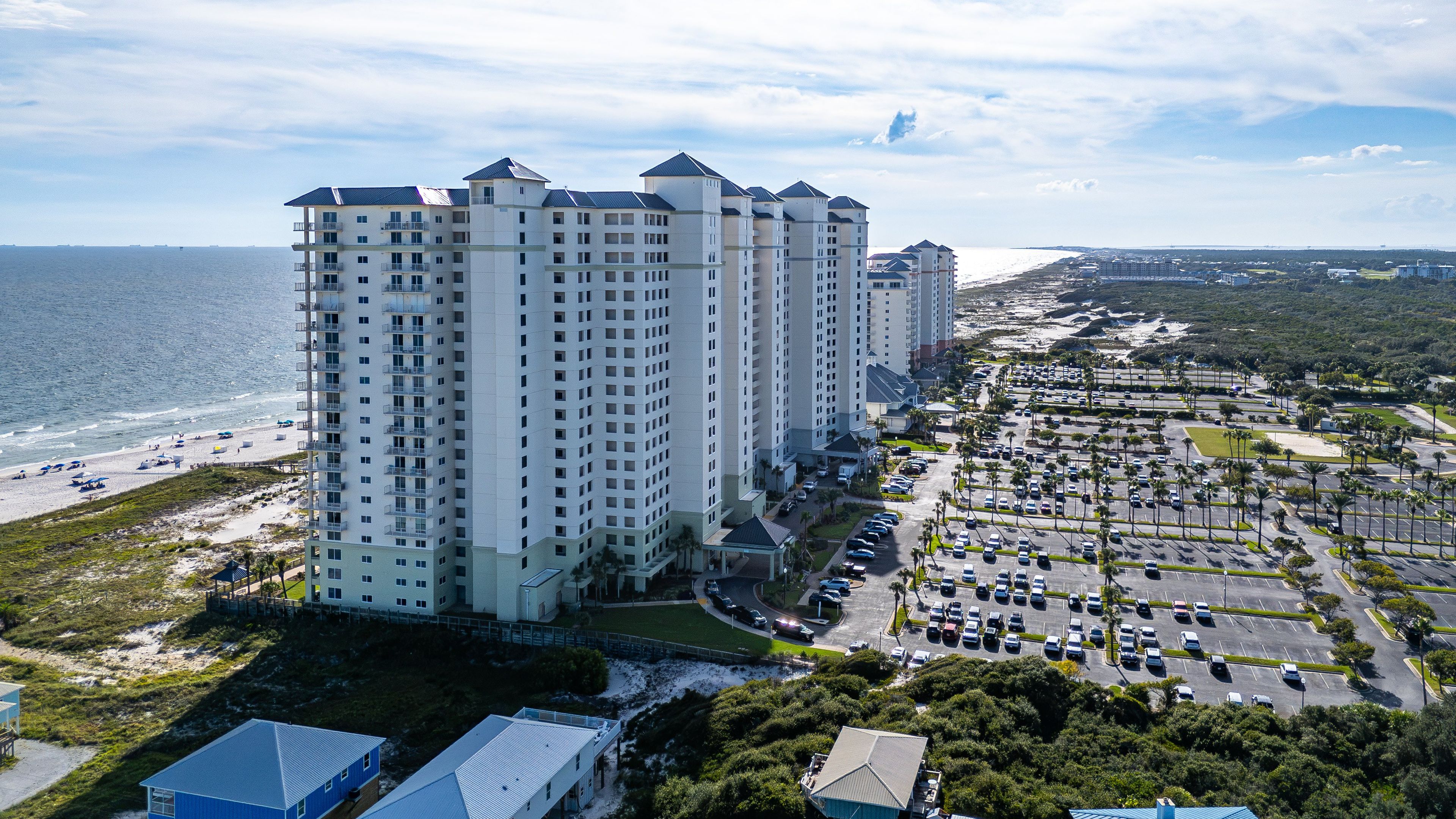 A high-rise hotel with multiple floors, a parking lot, and a beach in the background.