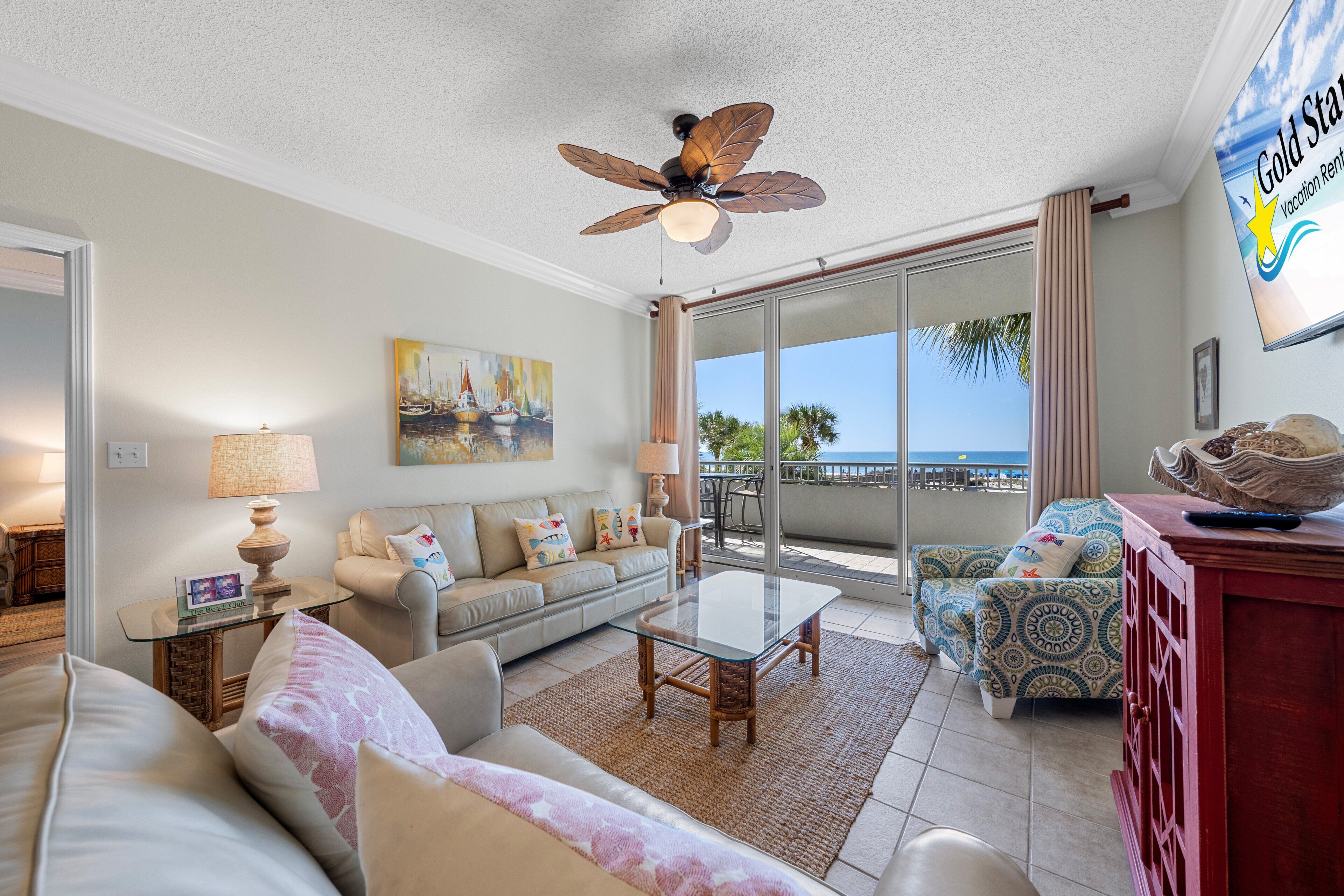 A living room with a ceiling fan, a glass table, a sofa, a patterned armchair, and a TV on the wall.