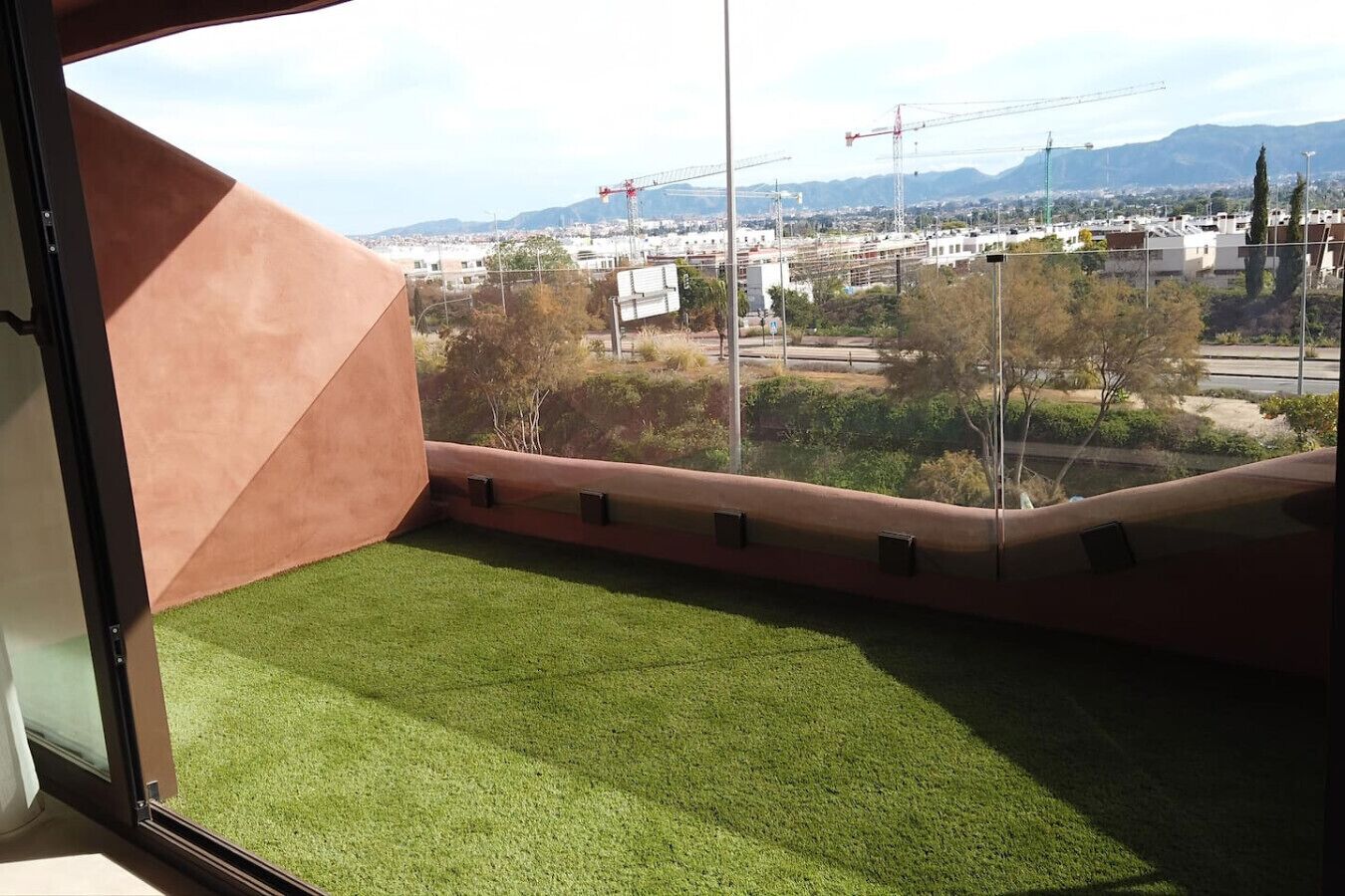 A balcony with artificial grass, a view of a cityscape with construction cranes, and distant mountains.