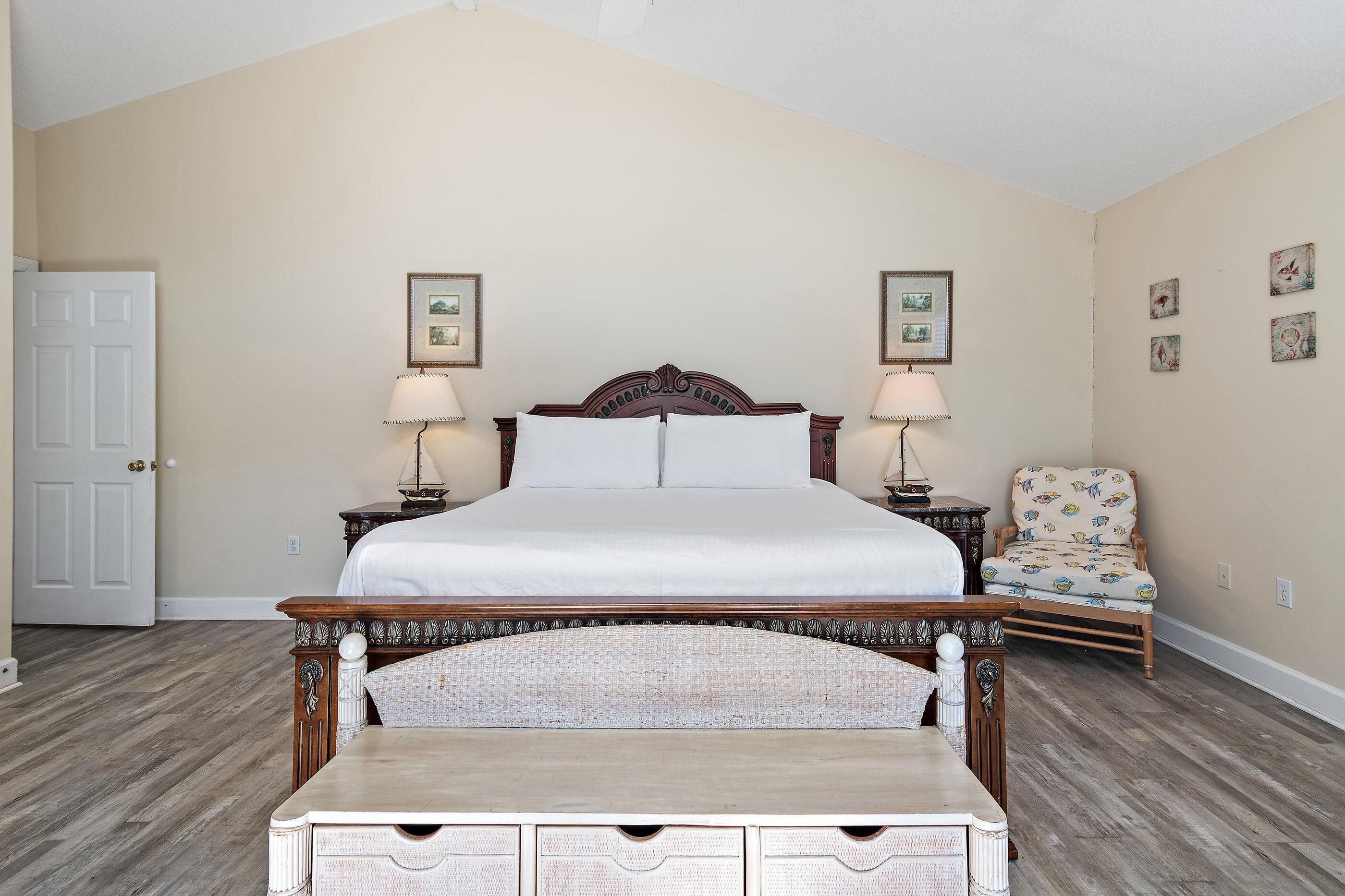 A bedroom with a large bed, two bedside tables with lamps, a chair with a floral pattern, and a wooden dresser with drawers.