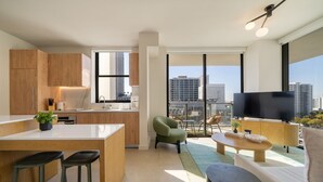 Eine moderne Wohnung mit Küche, Essbereich und Wohnzimmer. Sie verfügt über große Fenster mit Blick auf die Stadt, einen Flachbildfernseher und einen kleinen Balkon mit Sitzgelegenheit.