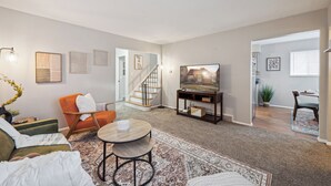 Una sala de estar moderna con sofá, sillón, mesa de centro y televisor sobre un mueble. Una escalera lleva a otra habitación.
