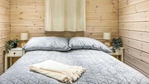 Ein Schlafzimmer mit einer holzverkleideten Wand, einem Bett mit gesteppter, grauer Bettdecke, zwei Nachttischen mit Lampen und einem Fenster mit Vorhängen.