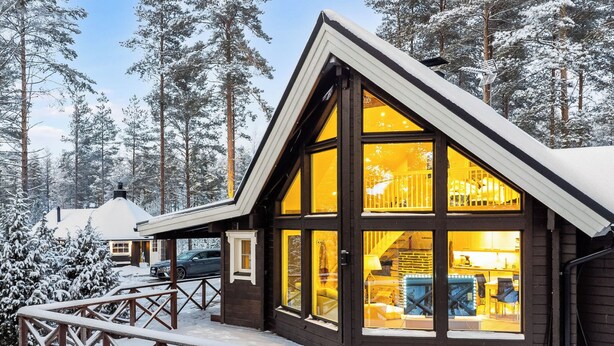 Eine Holzhütte mit großen Fenstern, einer Holzterrasse und einem verschneiten Waldbereich im Hintergrund.