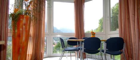 A dining area with a table and four chairs, large windows with curtains, and a tall vase with flowers.