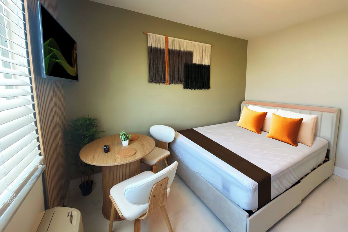 A hotel room with a bed, a small round table with a chair, a potted plant, and a wall-mounted TV.