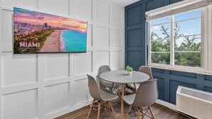 Ein modernes Zimmer mit Fernseher, der einen Sonnenuntergang am Miami Beach zeigt, einem runden Tisch mit Stühlen, einem Fenster mit Blick auf Bäume und einer weißen Wand mit Paneelen.
