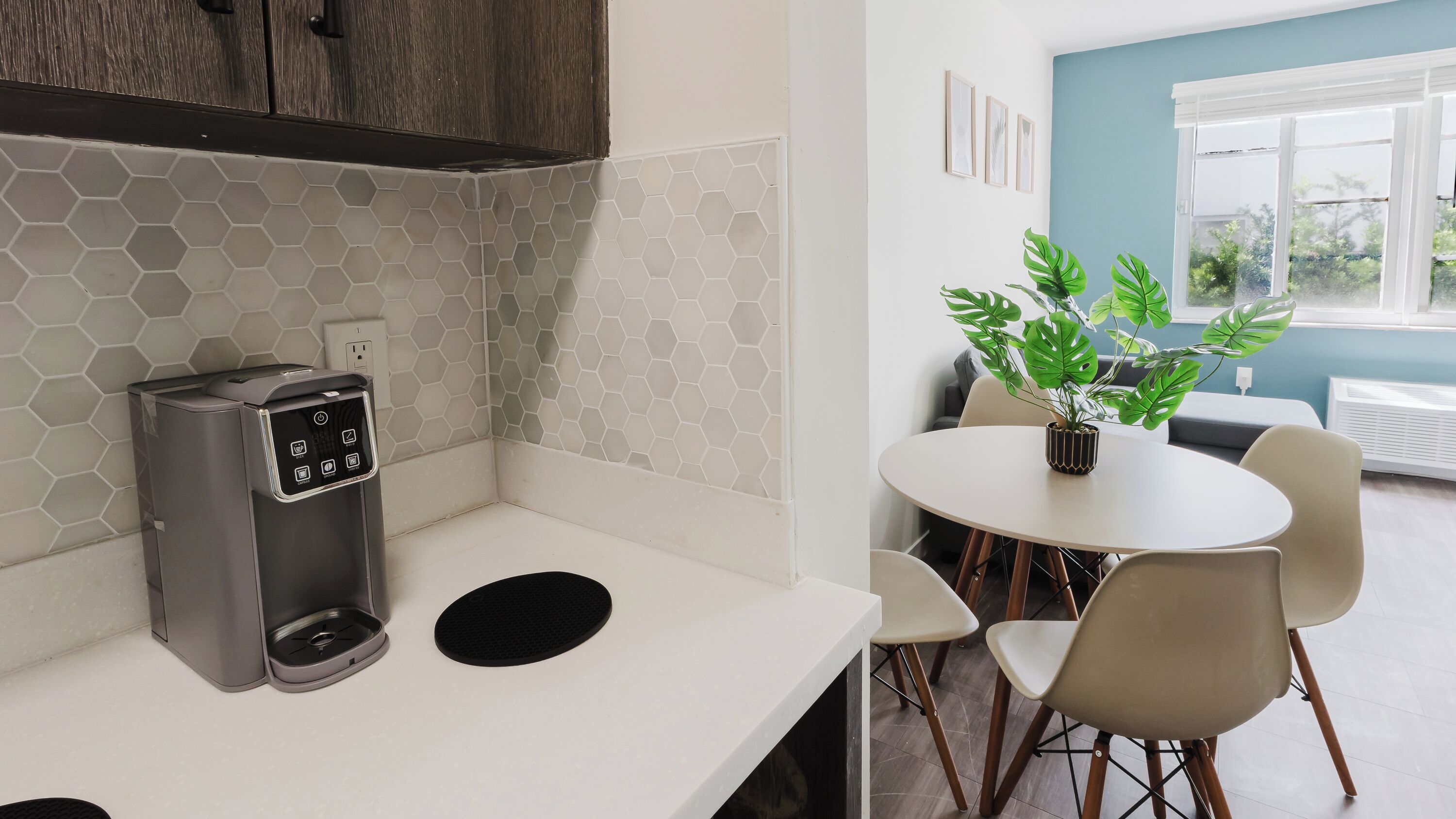 Una kitchenette con una cafetera, un salpicadero de azulejos hexagonales, una zona de comedor con mesa y sillas, y una planta en maceta.