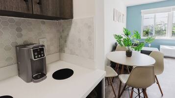 Una kitchenette con una cafetera, un salpicadero de azulejos hexagonales, una zona de comedor con mesa y sillas, y una planta en maceta.