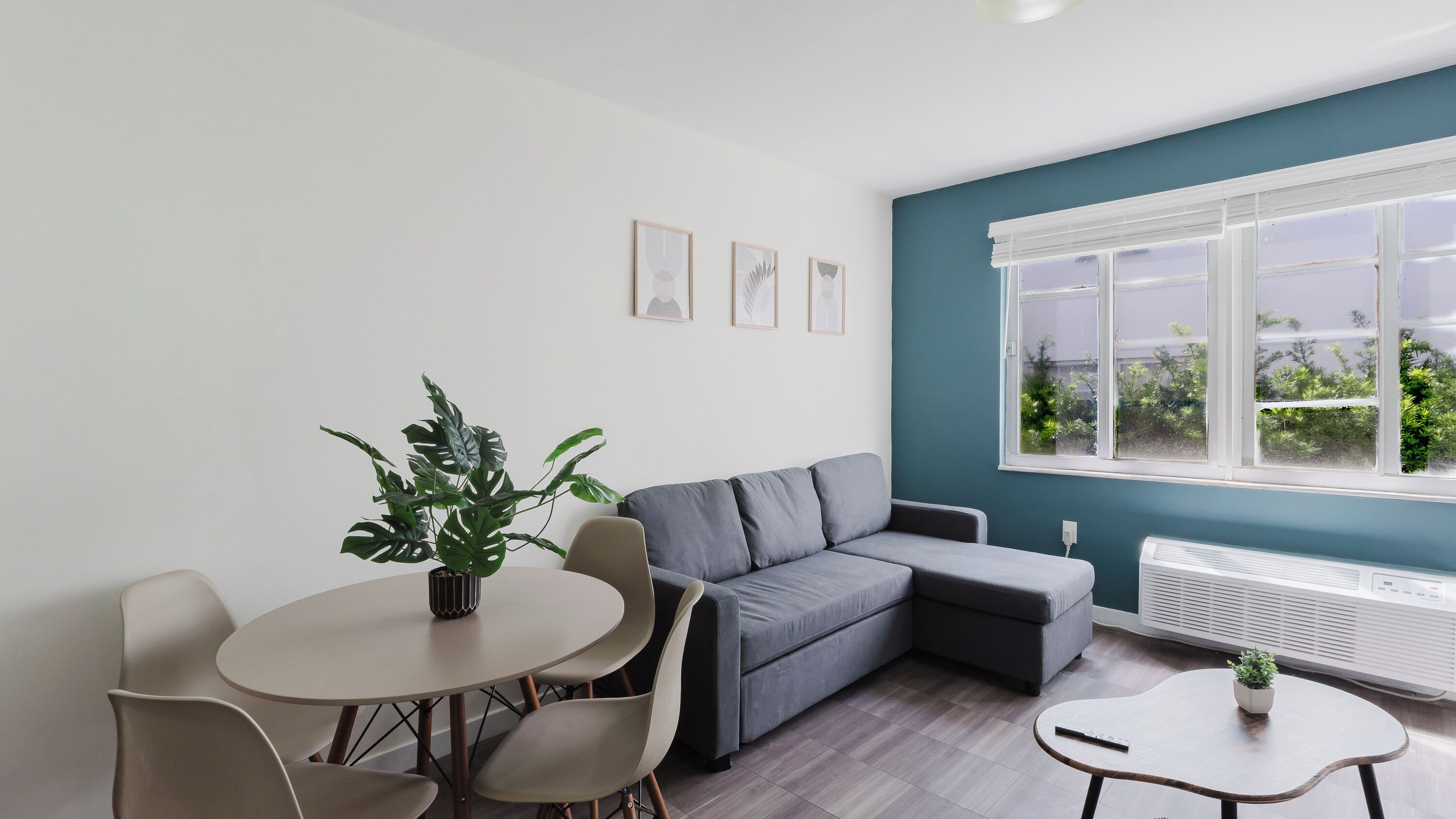 Un salon moderne avec un canapé gris, une table de salle à manger ronde entourée de chaises, une plante en pot et une fenêtre donnant sur un paysage verdoyant.