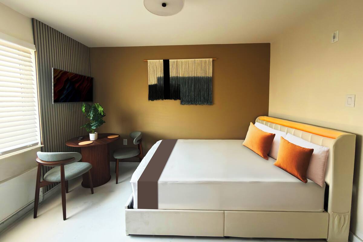 A modern hotel room with a large bed, a small round table, a chair, a potted plant, and a television on the wall.