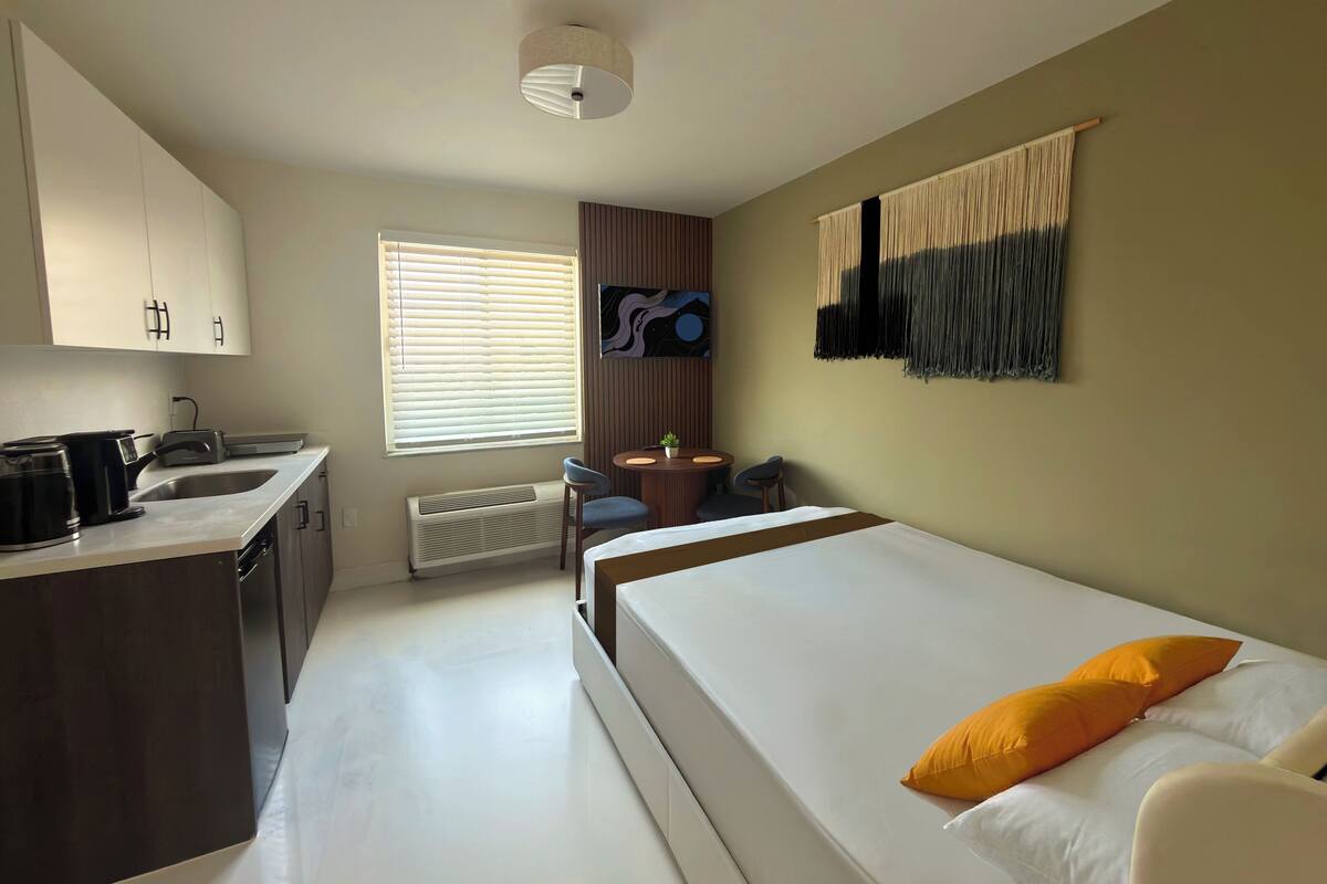A compact hotel room with a kitchenette, a bed with two pillows, a small table with chairs, and a wall-mounted artwork.