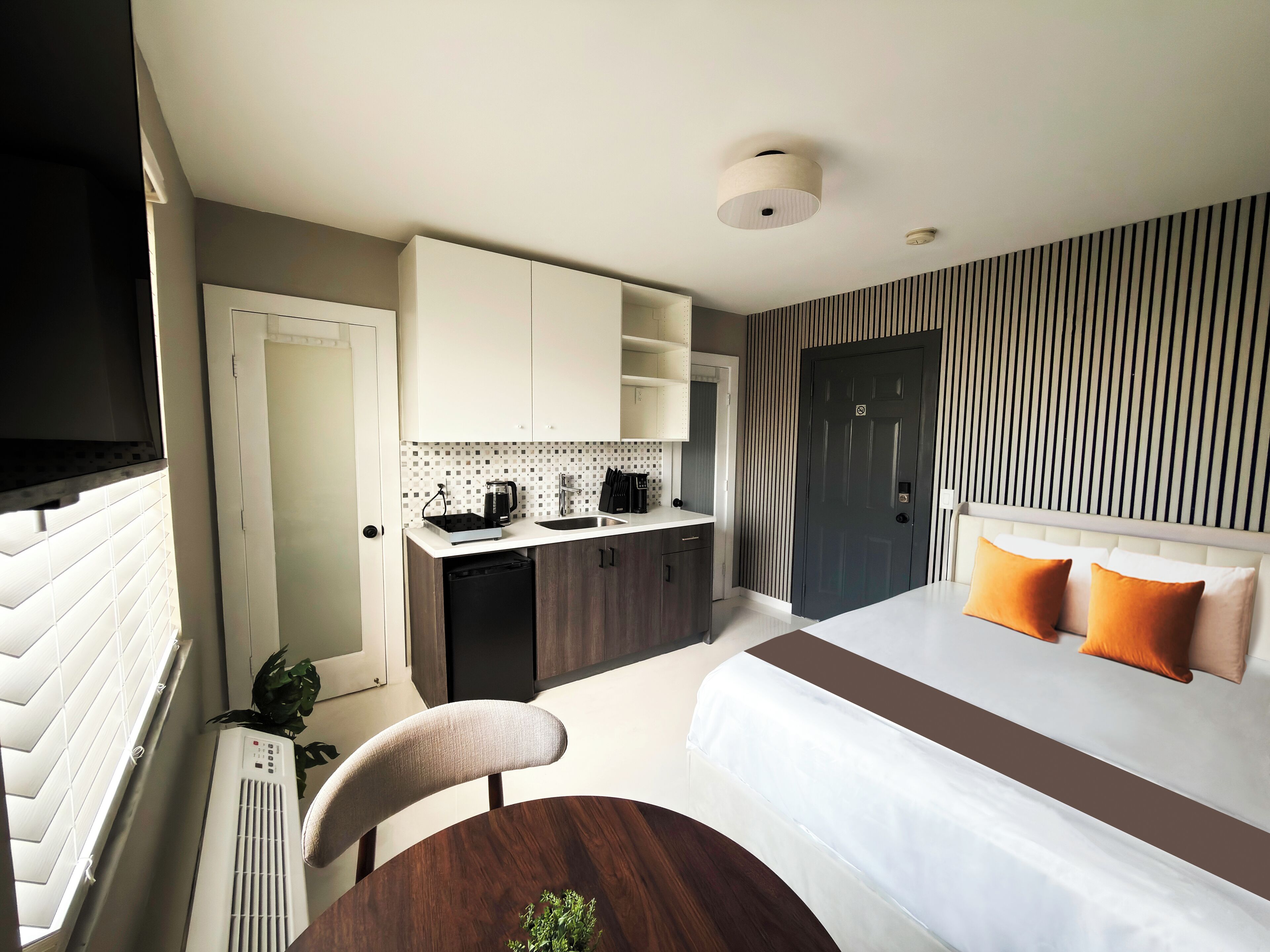 Una habitación de hotel compacta con kitchenette, una cama con almohadas anaranjadas, una mesa de comedor y un ventanal grande con persianas.