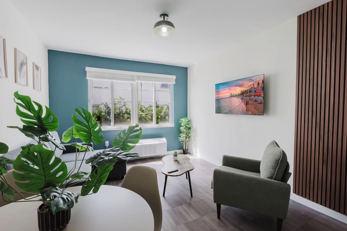 A modern living room with a large window, a dining table, a green armchair, and a potted plant.
