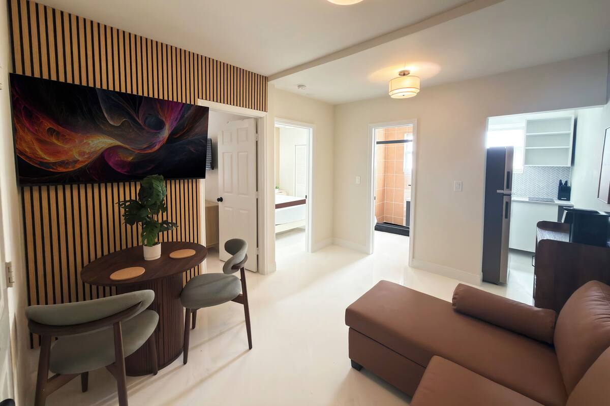 A modern living room with a brown sofa, a dining area with a wooden table and chairs, a TV mounted on a wooden panel, and a kitchen in the background.