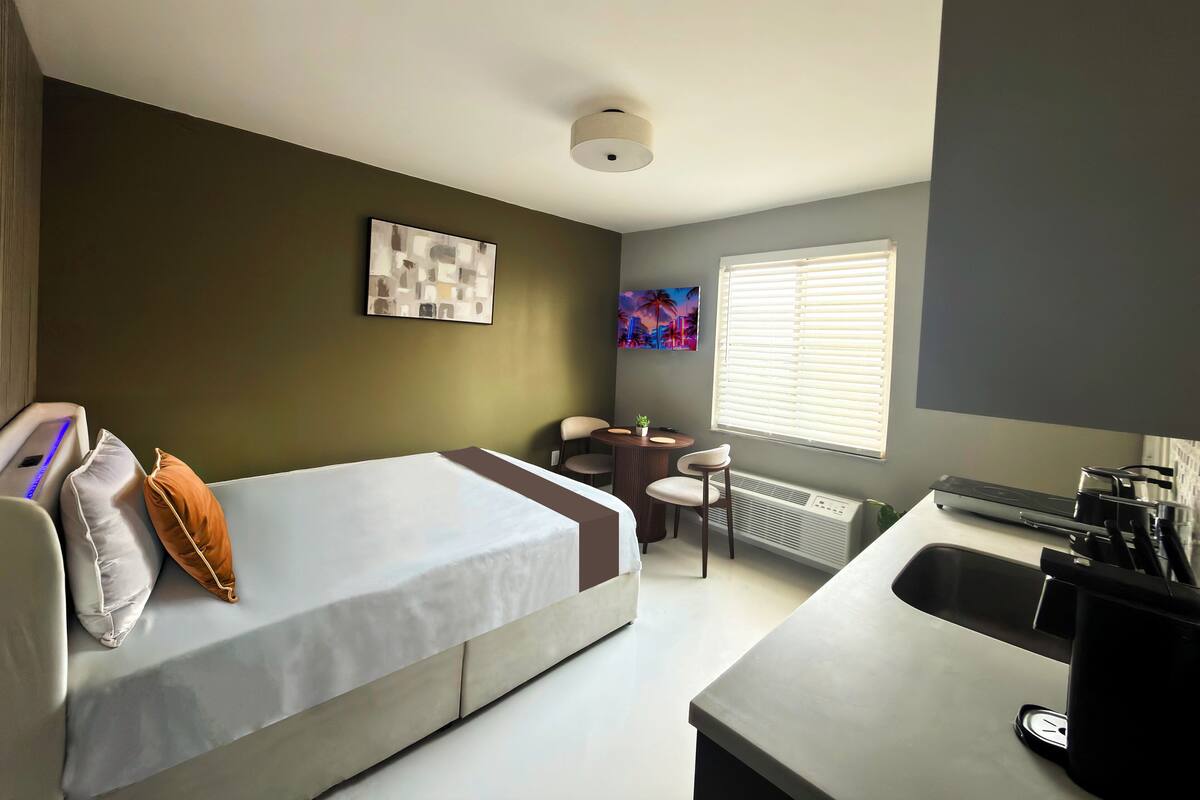 A compact hotel room with a bed, a small desk, a chair, a radiator, and a window with blinds.