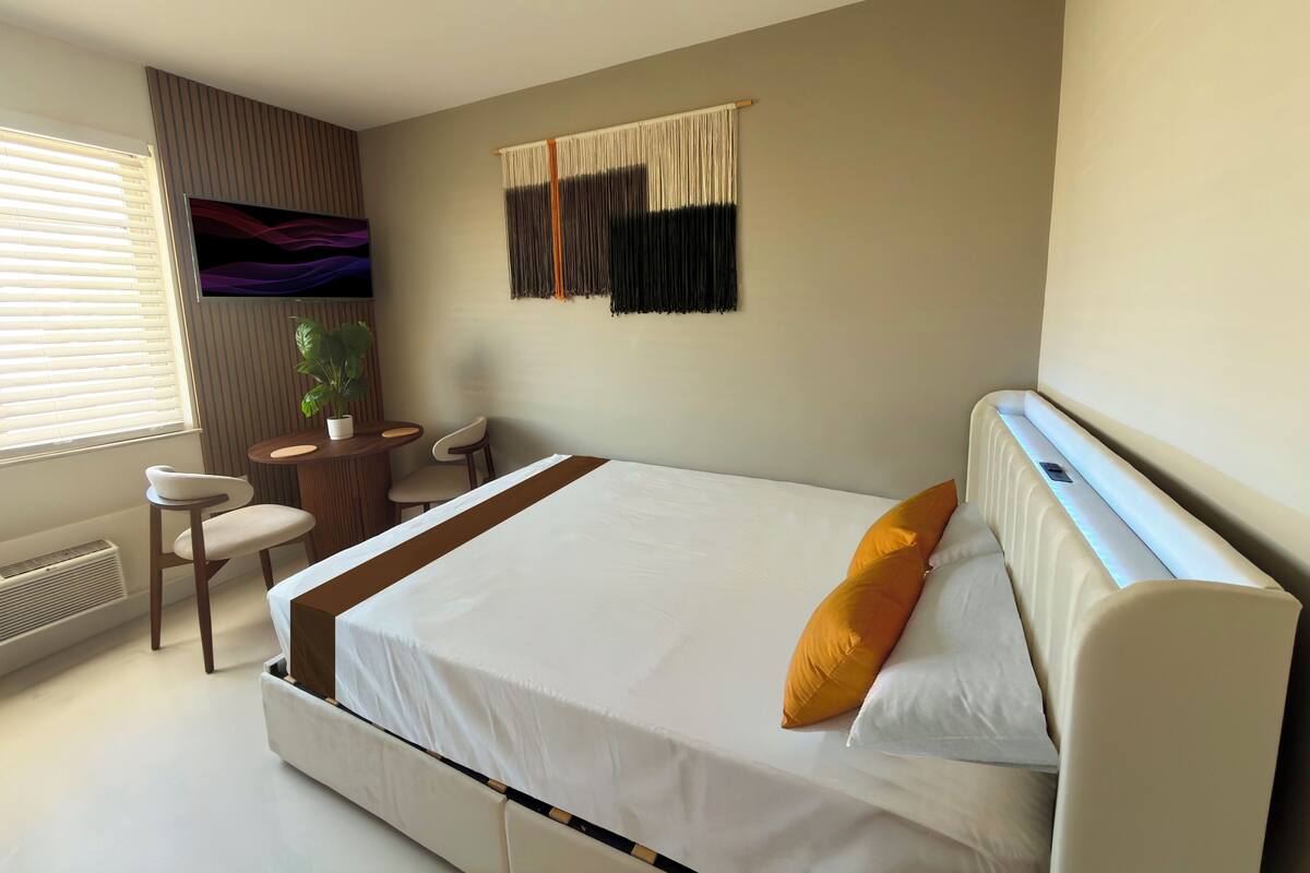 A modern hotel room with a large bed, a small desk, a chair, a potted plant, and a wall-mounted artwork.