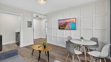 Ein modernes Wohnzimmer mit einem Flachbildfernseher, einem runden Esstisch, grauen Stühlen und einem kleinen Blumentopf.