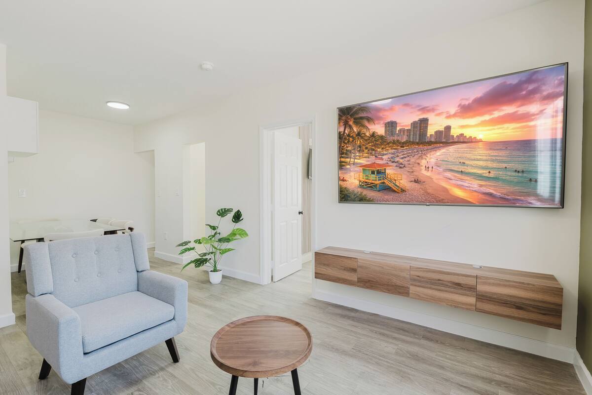 A modern living room with a light blue armchair, a round wooden table, a large beach mural, a potted plant, and a built-in wooden bench.