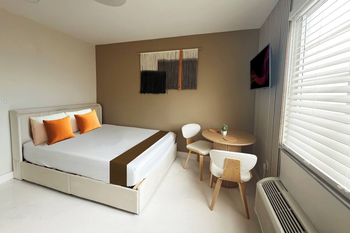 A modern hotel room with a bed, a small round table with chairs, a wall-mounted TV, and a window with blinds.