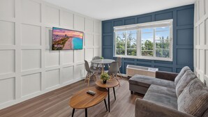 Ein modernes Wohnzimmer mit Flachbildfernseher, Esstisch, Stühlen, einer Couch und einem großen Fenster mit Blick auf Grünflächen.