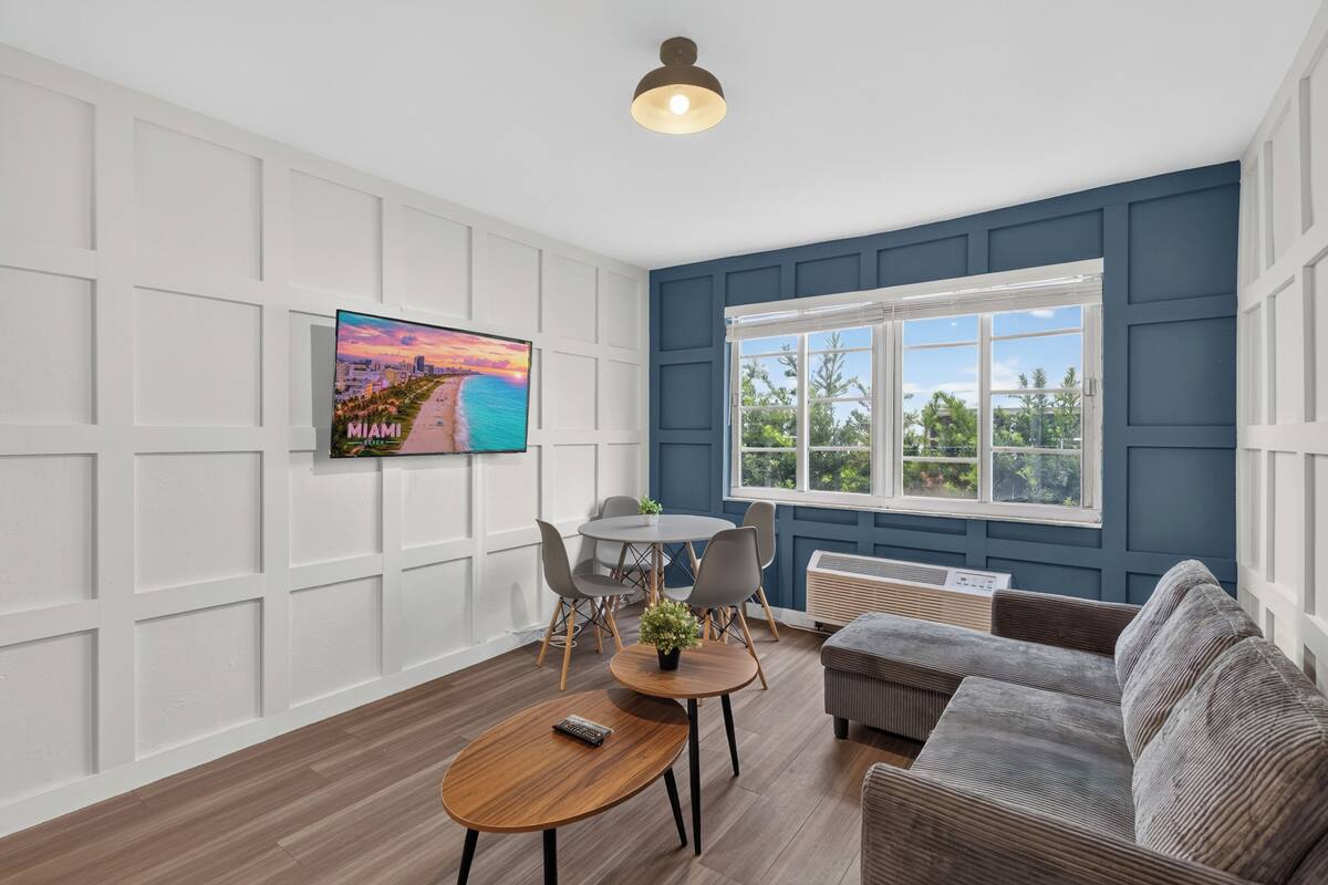 A modern living room with a flat-screen TV, a dining table, chairs, a sofa, and a large window with a view of greenery.