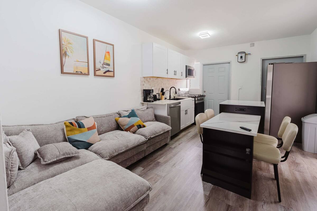 A modern living room with a sofa, a dining table with chairs, a kitchen area with cabinets and appliances, and two framed pictures on the wall.