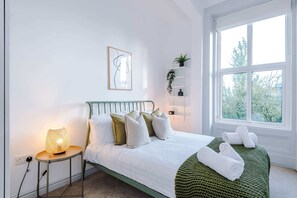 A bedroom with a bed, bedside table, lamp, wall art, shelf with plants, and a window. - 6BDR House | Sleeps 16 | Group Friendly (Manchester)
