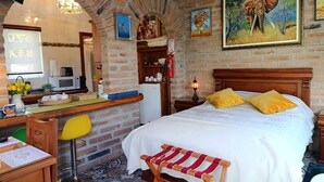 A cozy room with a brick wall, a bed with yellow pillows, a wooden bench, a desk with a chair, and a kitchen area.