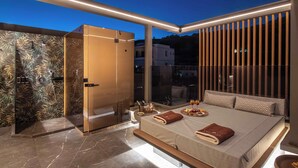 A modern bedroom with a glass shower, a bed with pillows, and a view of the city at dusk. - Lapis Nera Villas  (Viannos)