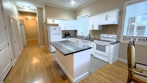 A modern kitchen with white cabinetry, a central island, and a dining area with a sign saying "let's eat". - Modern 2BR Walk-Out Suite | 2 Baths | Free Parking (Kelowna)
