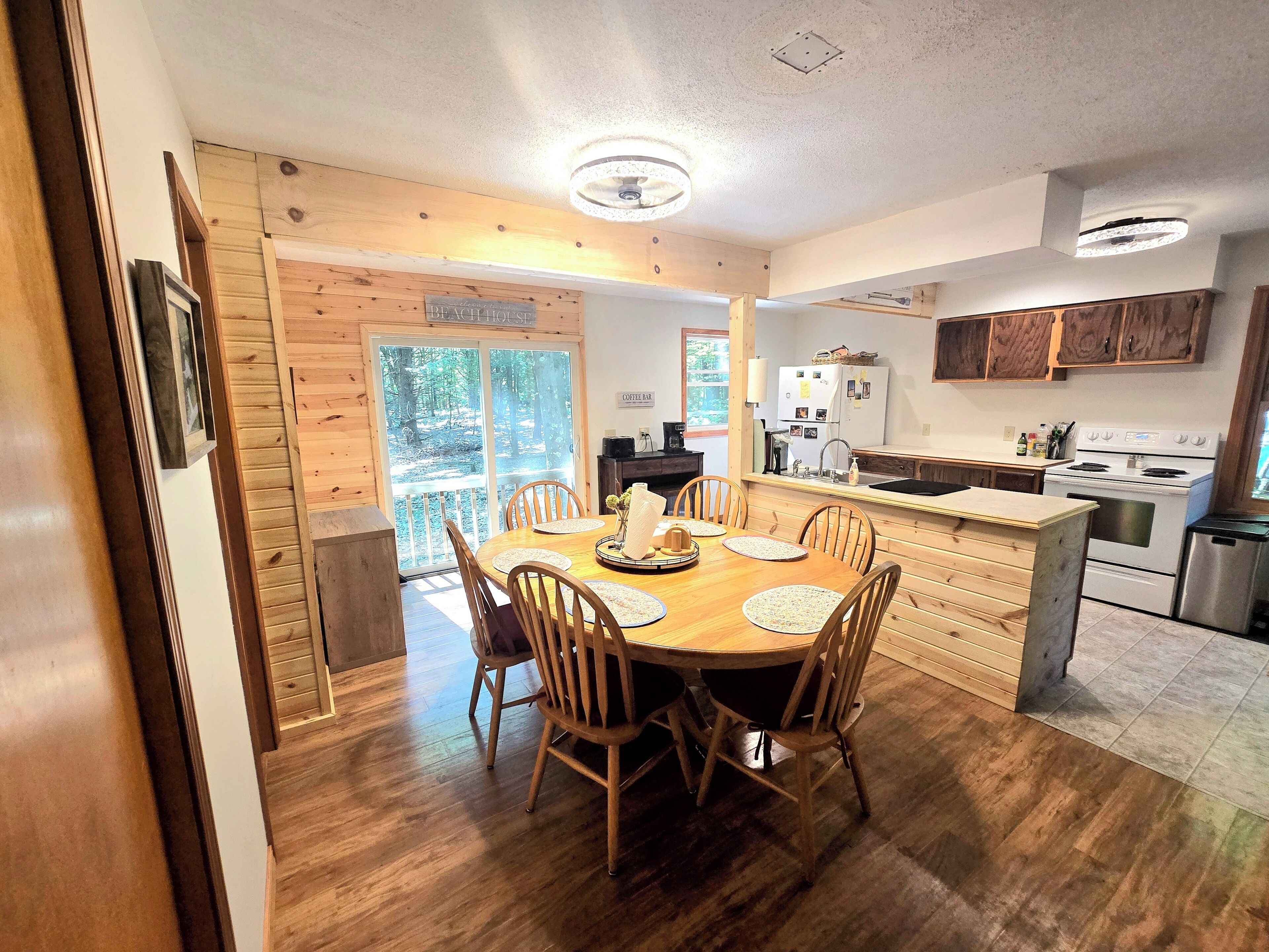 A wooden dining table with chairs, a kitchen with a stove and refrigerator, and a sliding glass door leading to a forested area.