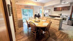 A wooden dining table with chairs, a kitchen with a stove and refrigerator, and a sliding glass door leading to a forested area. - NEW LISTING 15% OFF 5BR Lake Michigan Cottage Private Retreat • Family Friendly (Pentwater)