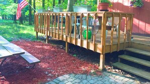 A wooden deck with a picnic table, surrounded by a red mulch bed, a red building, and a forested area with an American flag. - NEW LISTING 15% OFF 5BR Lake Michigan Cottage Private Retreat • Family Friendly (Pentwater)