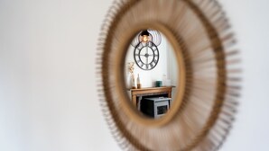 Un miroir avec un cadre circulaire reflétant une horloge murale, une petite étagère ornée d’objets décoratifs et une cheminée.