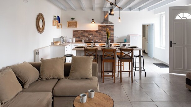 Un salon moderne avec un canapé, une table basse et un coin repas avec des chaises. On aperçoit, au fond, une cuisine avec un mur de fond en briques et des appareils électroménagers blancs.