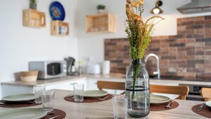 Un coin repas avec une table en bois dressée pour quatre personnes, un vase contenant des fleurs séchées et, en arrière-plan, un mur recouvert de briques apparentes.