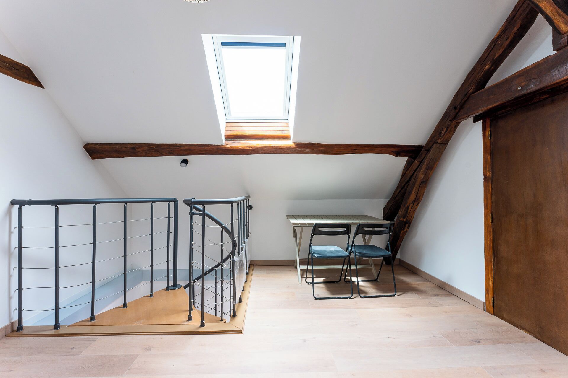 Une petite pièce mansardée avec un plafond en pente, un escalier en bois, une table avec deux chaises et une fenêtre.