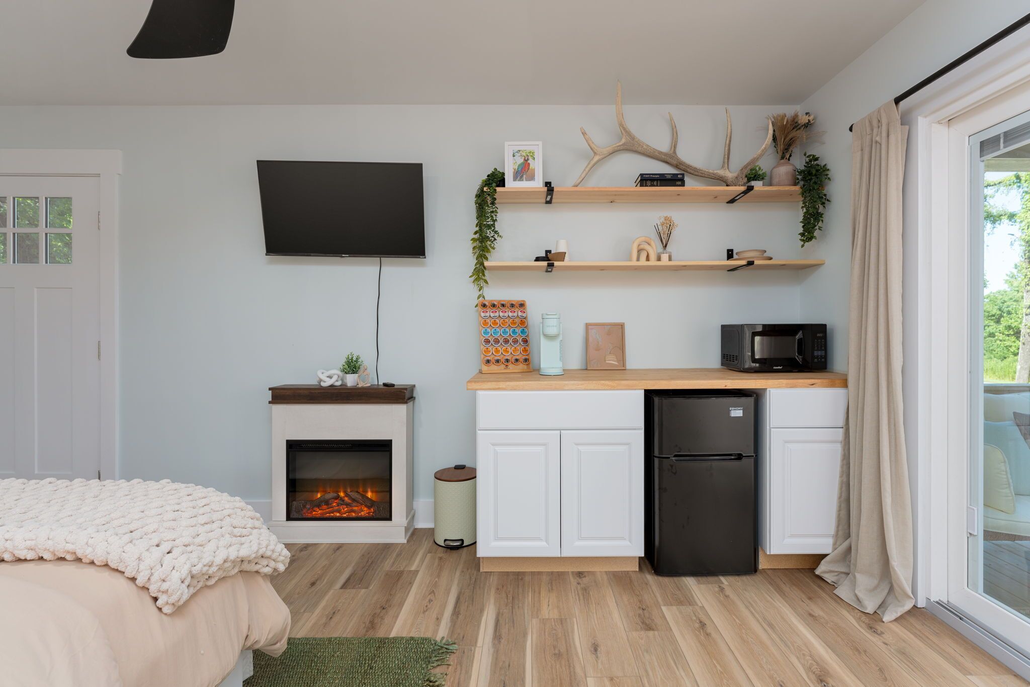 Een moderne slaapkamer met een open haard, een bed met een gebreide deken, een aan de muur bevestigde televisie, en een kitchenette met een koelkast, magnetron en houten planken.