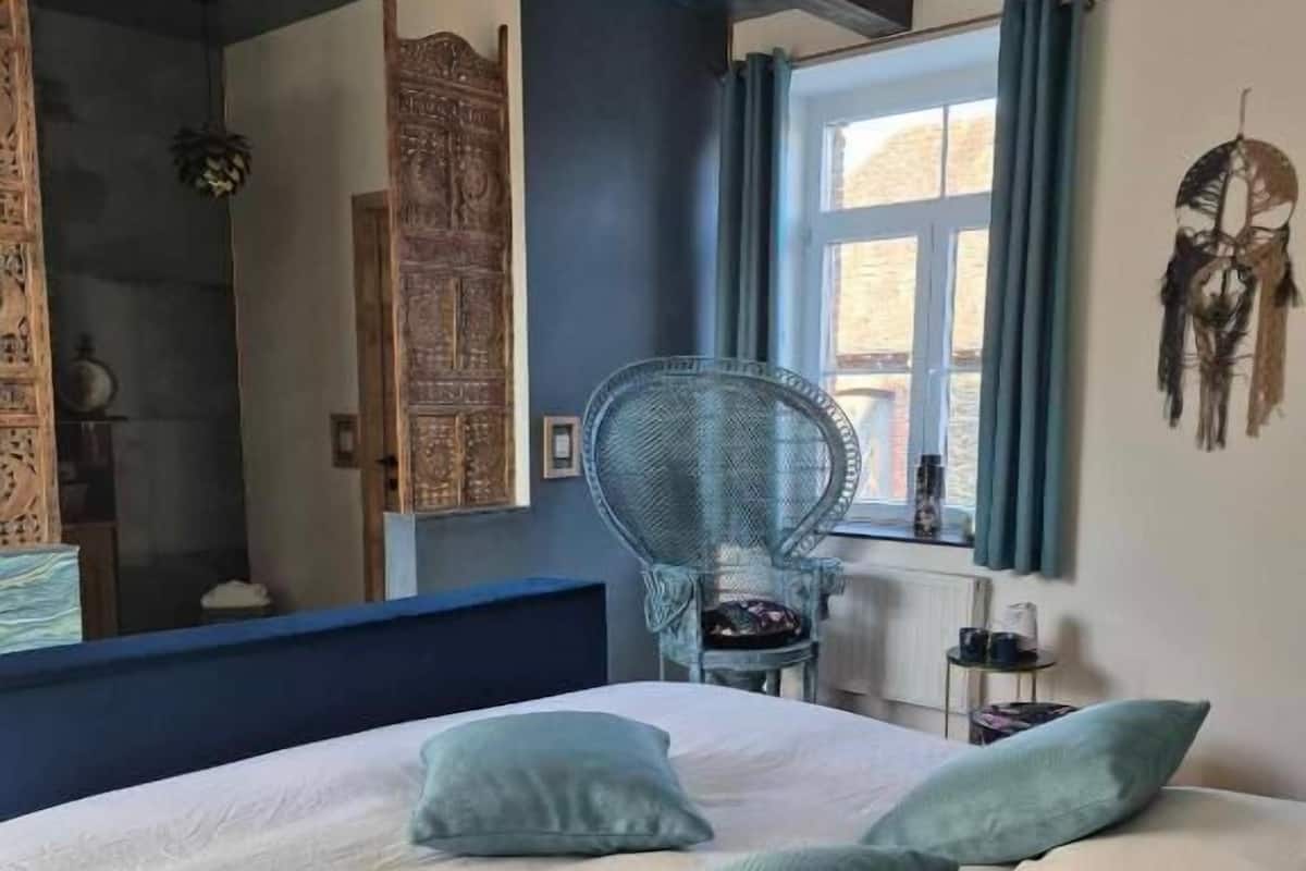 A bedroom with a large bed, a chair, a window with curtains, and a wall with a dreamcatcher.