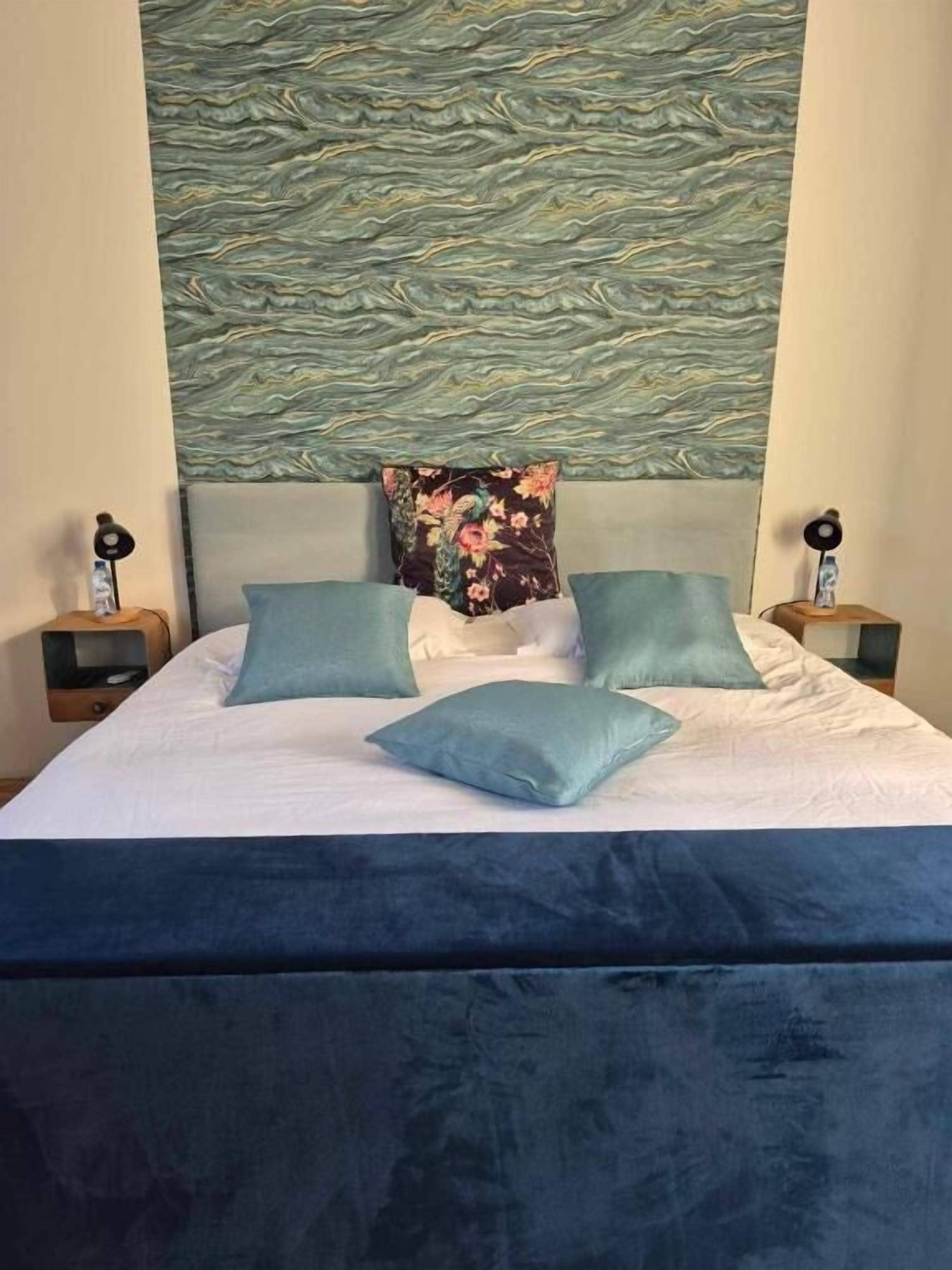 A bed with a dark blue headboard, white bedding, and multiple pillows in shades of blue and green. There are two bedside tables with lamps on either side of the bed.