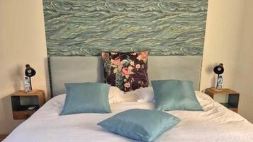 A bed with a dark blue headboard, white bedding, and multiple pillows in shades of blue and green. There are two bedside tables with lamps on either side of the bed.