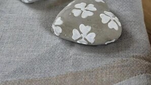 Dos piedras lisas con diseños de trébol blanco sobre una superficie de tela texturizada.