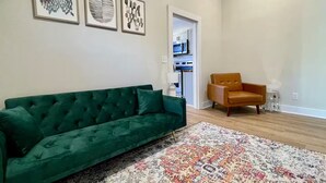 Ein Wohnzimmer mit einem grünen Sofa, einem gemusterten Teppich und einem gelben Sessel. An der Wand hängen drei gerahmte Kunstwerke.