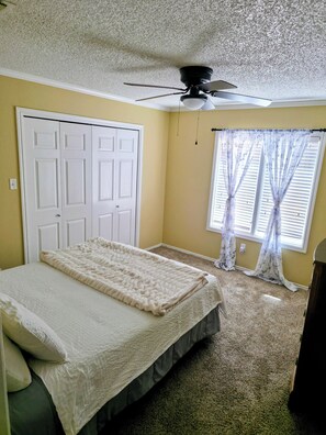 A bedroom with a bed, a ceiling fan, a window with sheer curtains, and a carpeted floor.