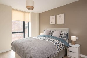 Ein Schlafzimmer mit einem großen Fenster, einem Bett mit gemusterten Betttextilien, zwei Wandbildern und einem Nachttisch mit Lampe.