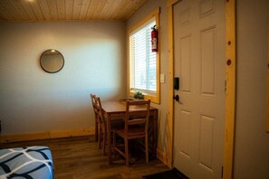 Une pièce avec une table et des chaises en bois, un miroir rond accroché au mur, et une porte dotée d’une fenêtre et de stores.