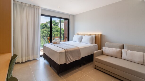 A modern hotel room with a large bed, a sofa, and a balcony with a view of greenery. - Modern Studio in Lapa With Pool and gym R1205 (Nova Iguaçu)
